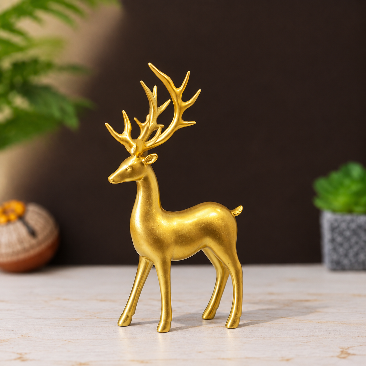 A shiny gold stag figurine with antlers standing on a light marble surface, with a dark brown background and blurred green plants in the scene.