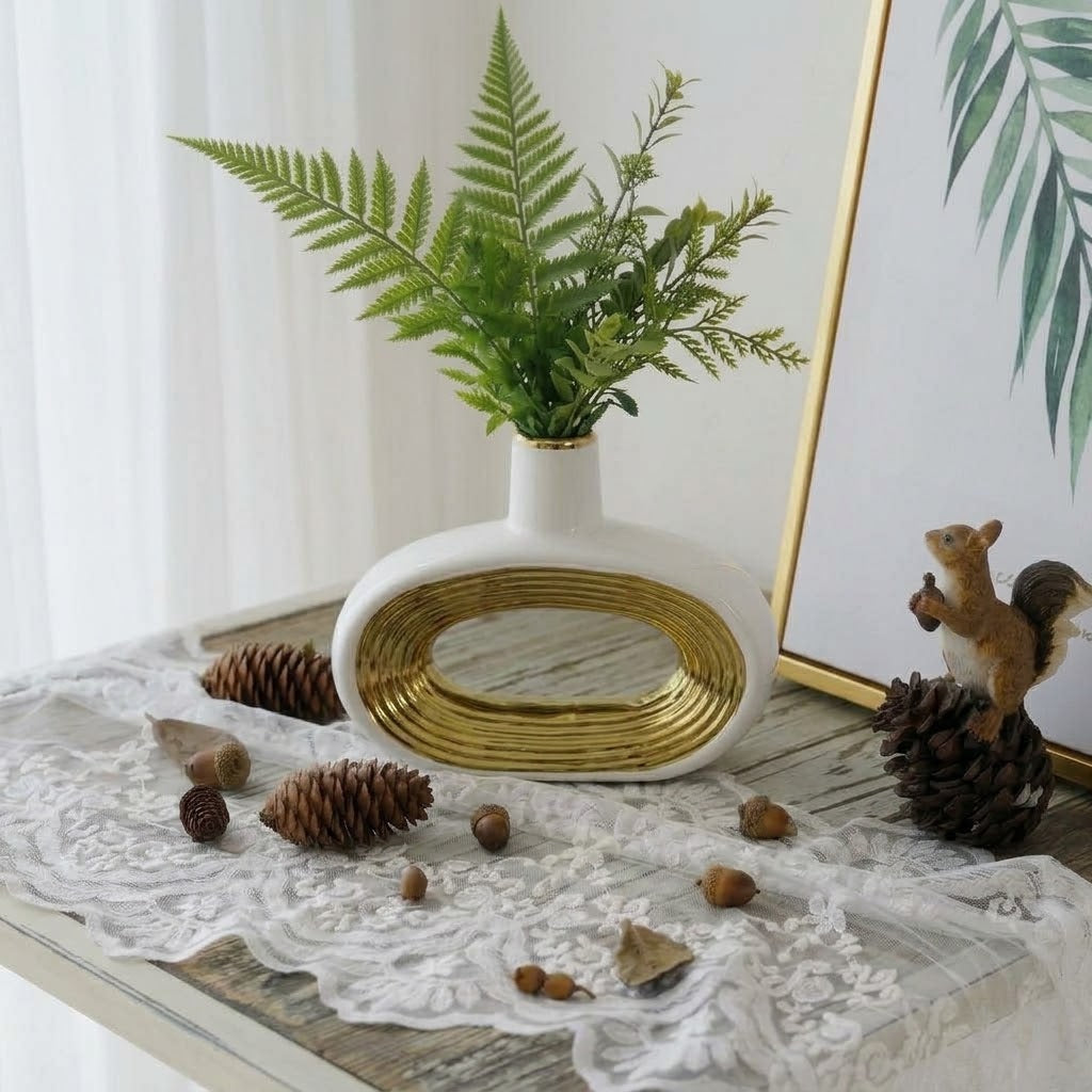 A white ceramic vase with a gold metallic oval accent and a small opening, filled with green fern-like foliage, styled on a lace-covered table with pinecones, acorns, and a framed print in a bright indoor setting.