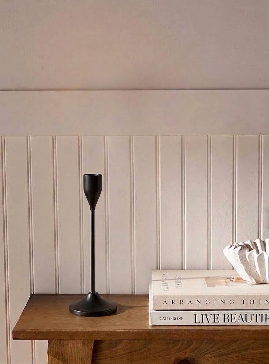 A matte black metal candlestick holder with a long stem and wide circular base, displayed on a wooden console table next to stacked books against a beige paneled wall.