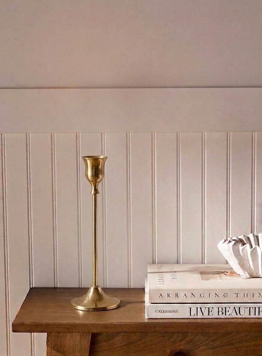 A brass candlestick holder stands on a wooden console table against a beige paneled wall, next to stacked books and a white decorative object.