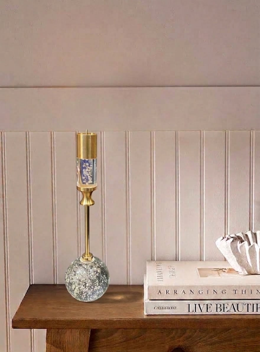 A decorative table lamp with a gold-finished metal stem and base, featuring a blue-and-white patterned glass upper section and a textured clear glass round base, displayed on a wooden shelf next to books against a paneled wall.