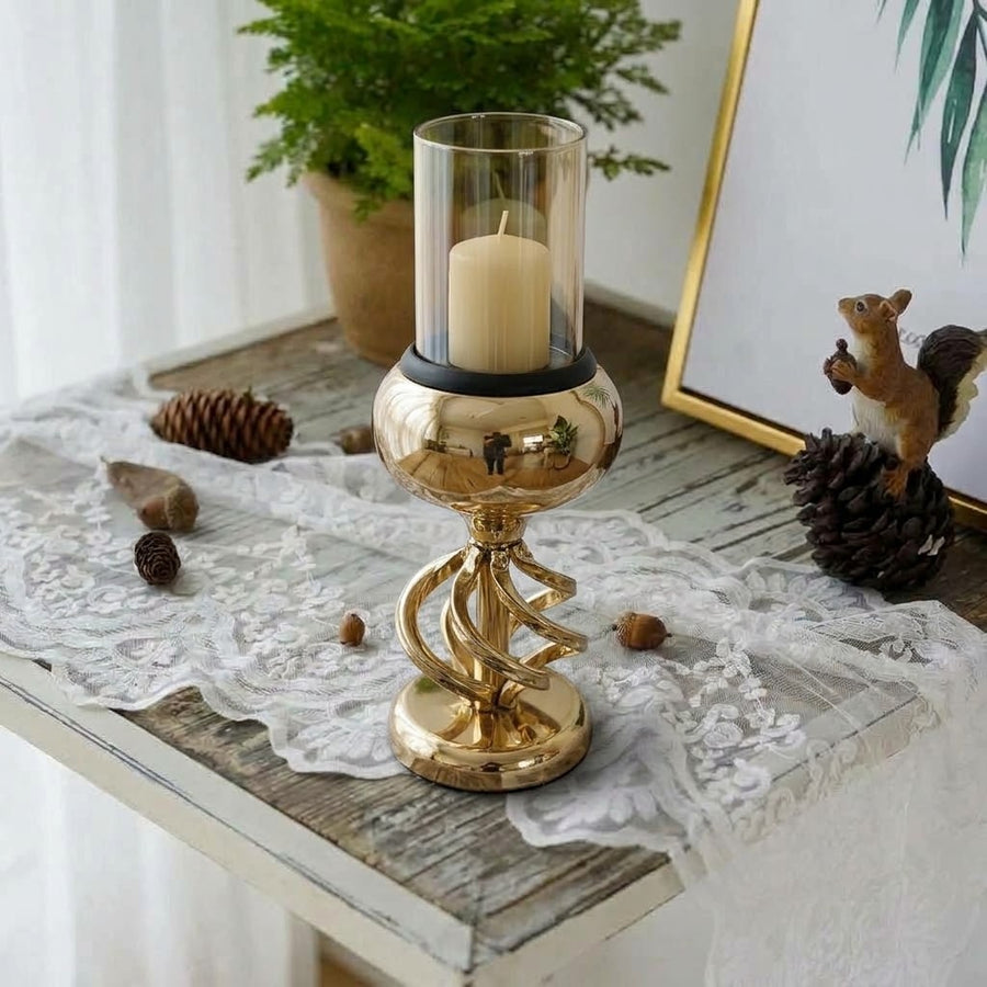 A gold-toned spiral pedestal candle holder with a clear glass cylinder and cream candle, styled on a rustic table with lace, pinecones, and a framed print.