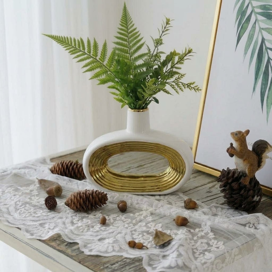 A white ceramic vase with a gold metallic oval accent and a small opening, filled with green fern-like foliage, styled on a lace-covered table with pinecones, acorns, and a framed print in a bright indoor setting.
