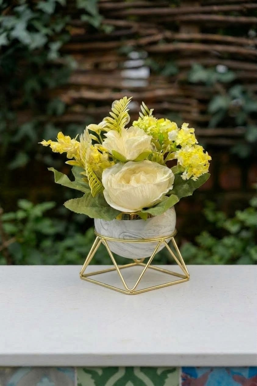 A floral arrangement with pale yellow flowers and green foliage in a small white marble-patterned vase on a gold geometric metal stand, placed on a white ledge with a blurred garden background.